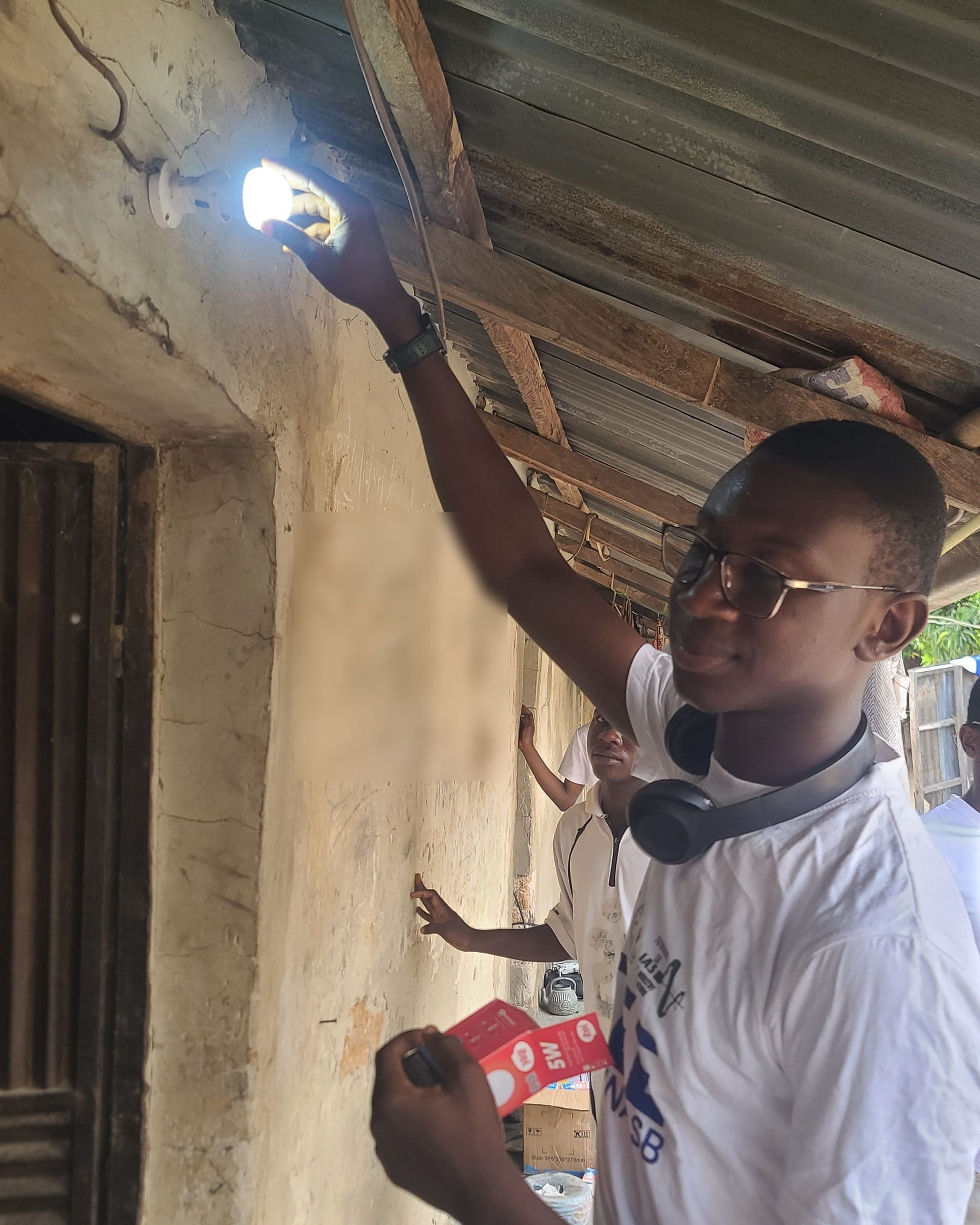 A smiling SolarPeer team member holds up a lit bulb under the edge of a roof, with the bulb's box in the other hand.