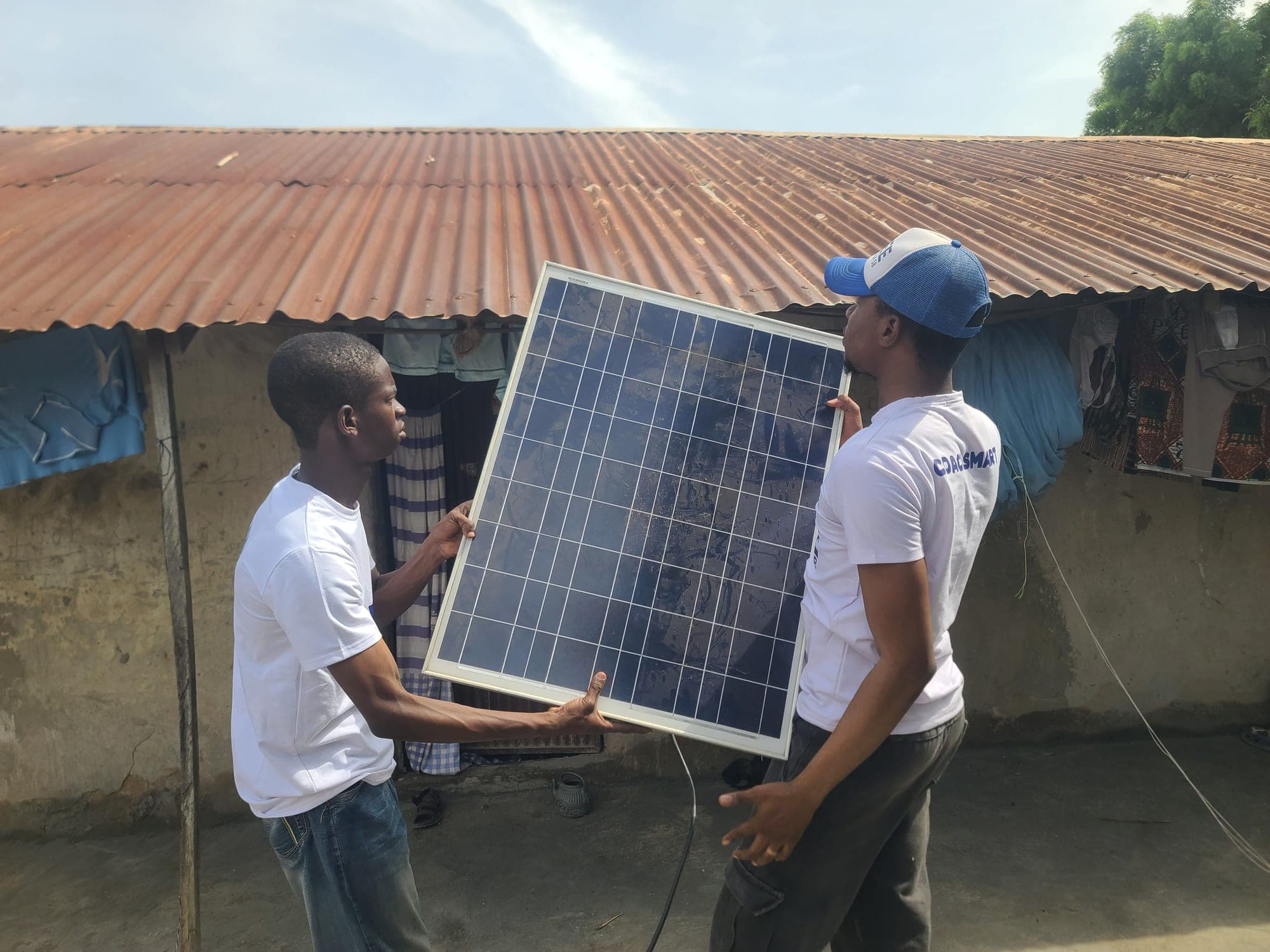 Two SolarPeer team members hold up a solar panel outside a house with a corrugated iron roof in Minna.