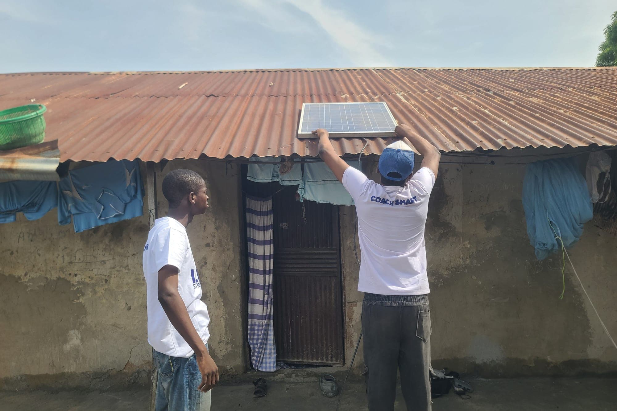 A SolarPeer team member fixes a small solar panel to the edge of a corrugated iron roof while a colleague watches.
